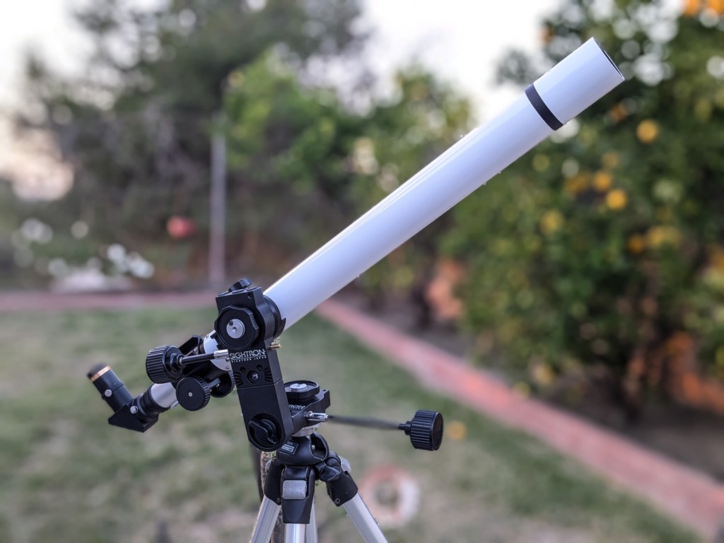 A white telescope mounted on a black tripod, positioned outdoors with blurred green trees in the background.