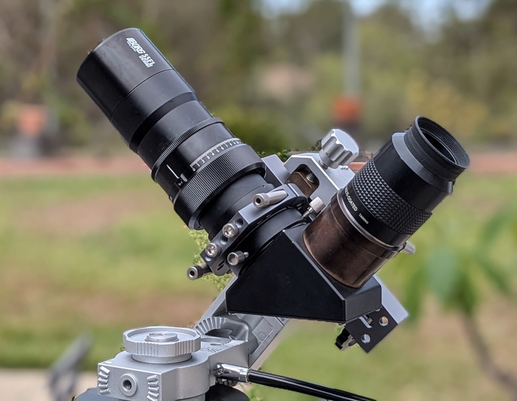 A close-up of a telescope attachment with two camera lenses mounted on a metal base, set against a blurred outdoor background.